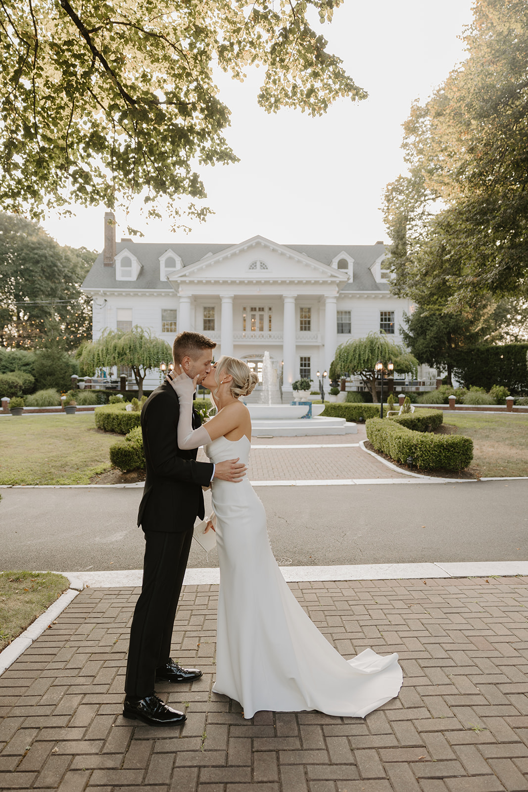 Couple shares a romantic kiss on a brick pathway with the grand white mansion and fountain behind them.